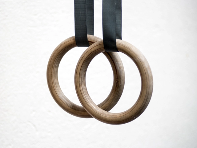 Blog 4 Wooden gymnastics rings hanging on black straps against a white background.